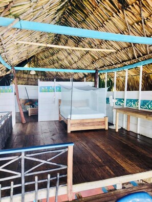 A thatched-roof structure with wooden flooring, a bed, a table, and a bench.