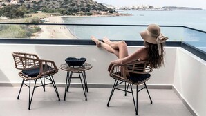 Ein Balkon mit Korbstühlen und einem kleinen Tisch, mit Blick auf einen Strand und eine hügelige Landschaft.