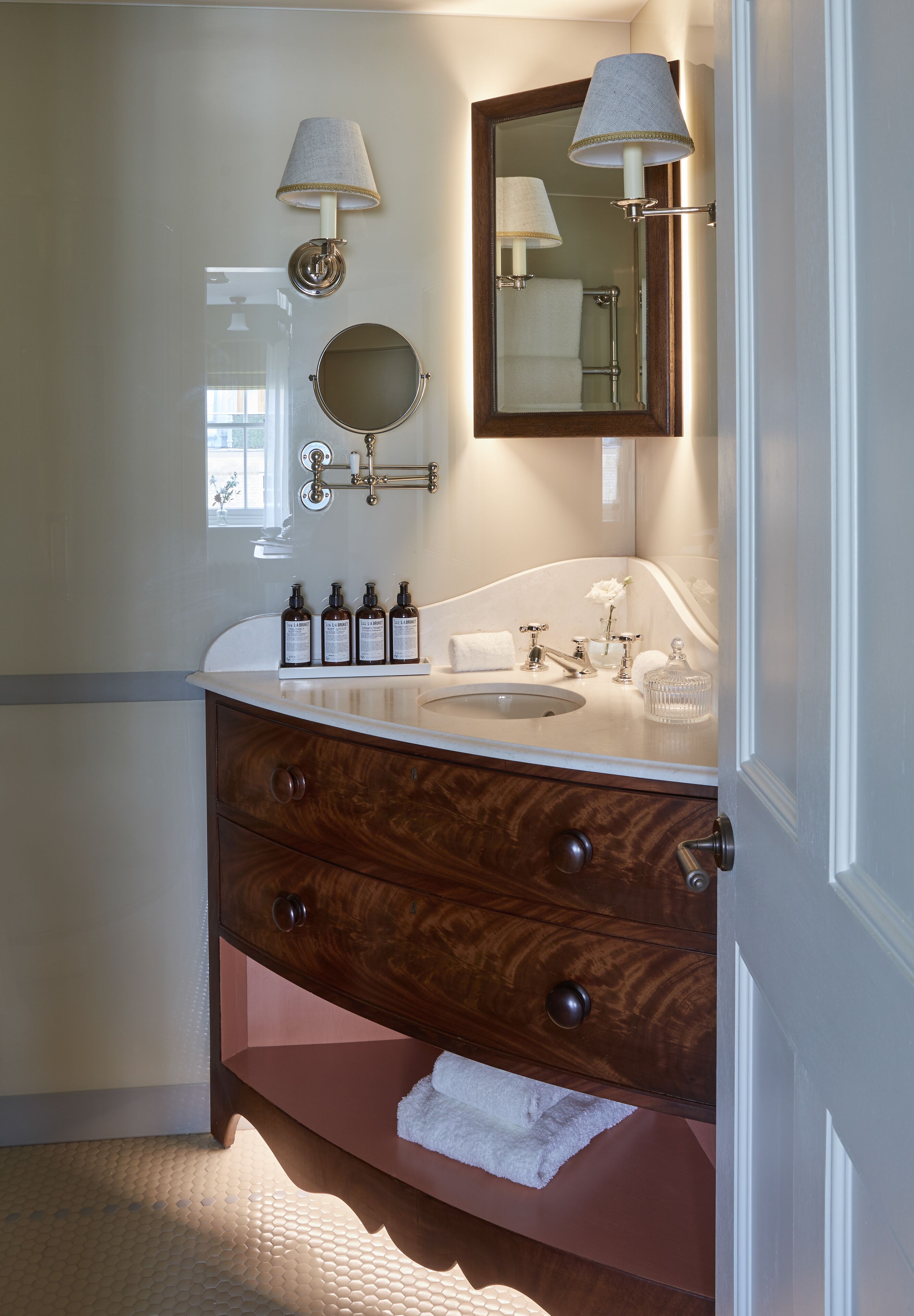 A bathroom with a wooden vanity, a round mirror, two wall-mounted lamps, a sink, and a window providing natural light.