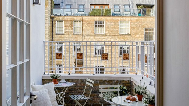Un balcón con vistas a un edificio de ladrillo, mobiliario de metal blanco y una pequeña mesa con una planta y un plato.
