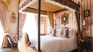 Una cama con dosel sobre cuatro columnas, con estructura de madera y cubrecama blanco, frente a una pared a rayas rosa y blanca con apliques decorativos y un espejo enmarcado.