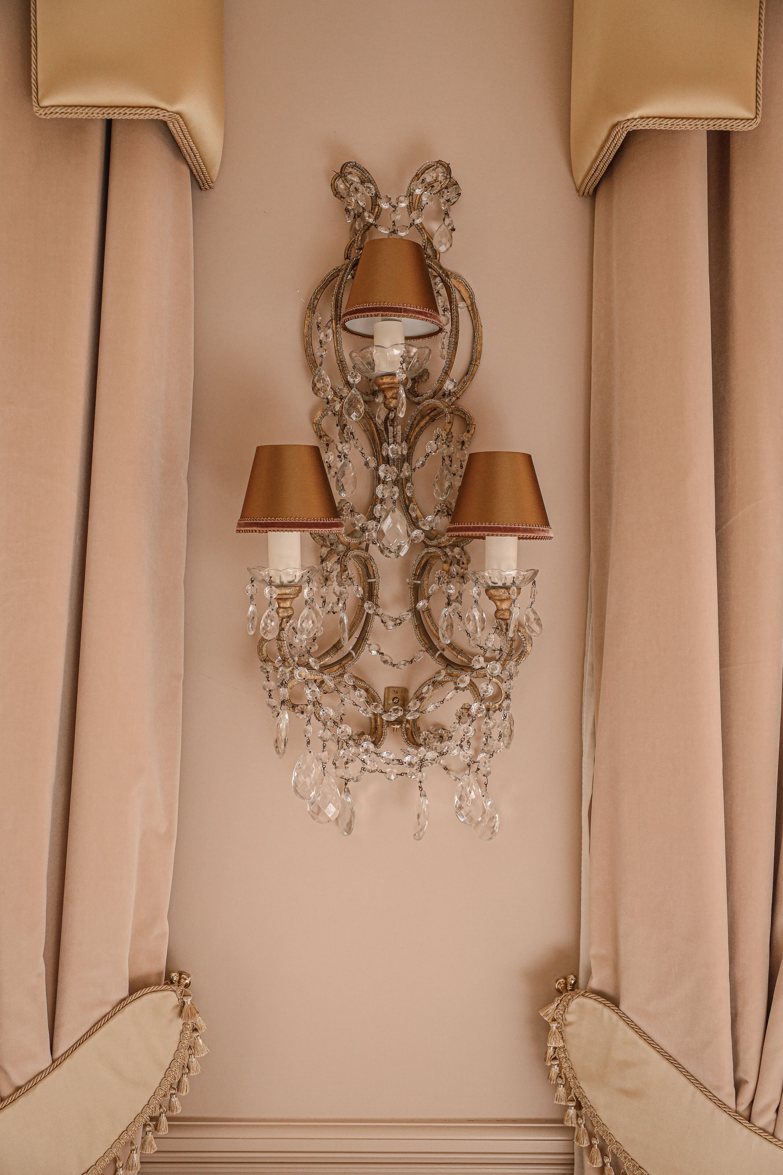 A wall-mounted chandelier with three lamps, set against a beige wall with draped curtains.