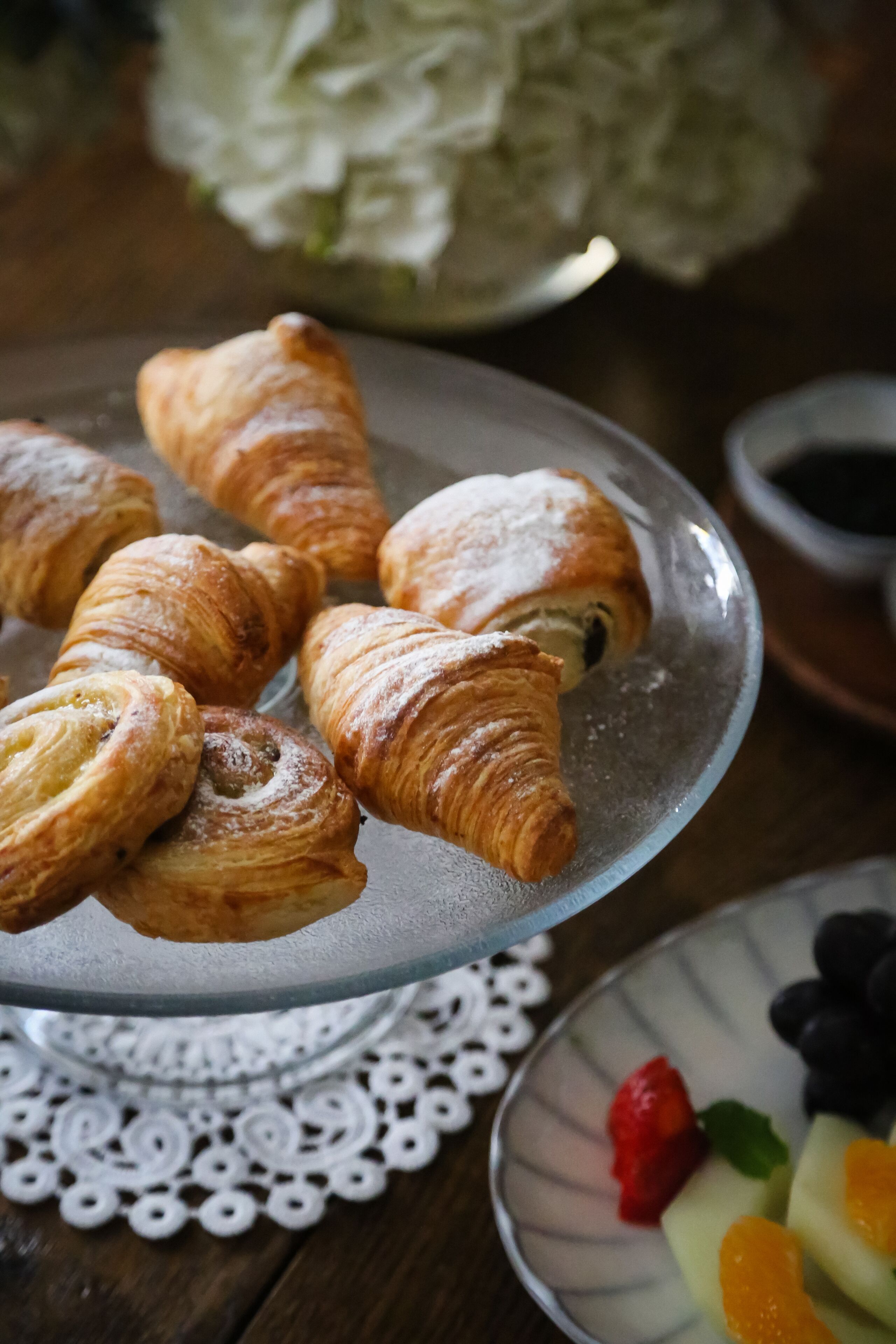 Un plato de croissants sobre un soporte de vidrio con un mantel de encaje debajo.