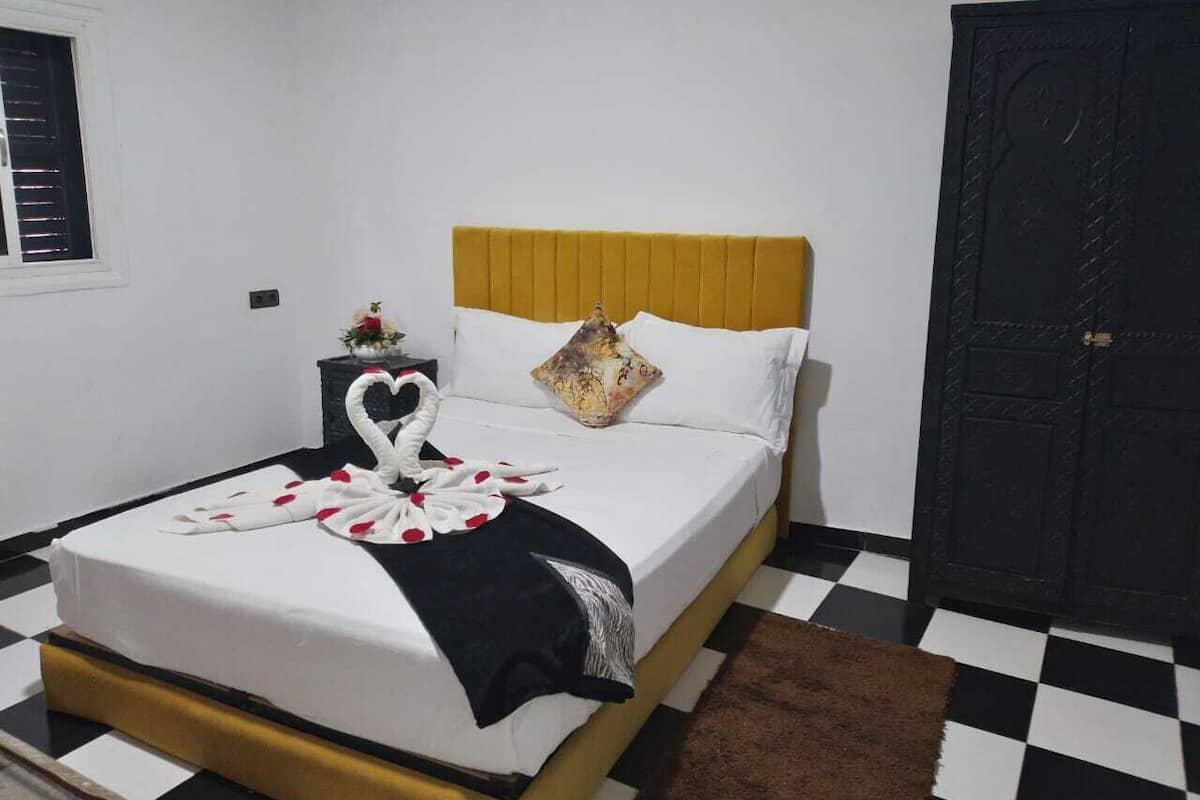 A neatly made bed with a swan towel arrangement, a patterned pillow, and a black and white checkered floor.
