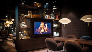 A cozy bar area with a television displaying a fire, plush chairs, and a wooden table.