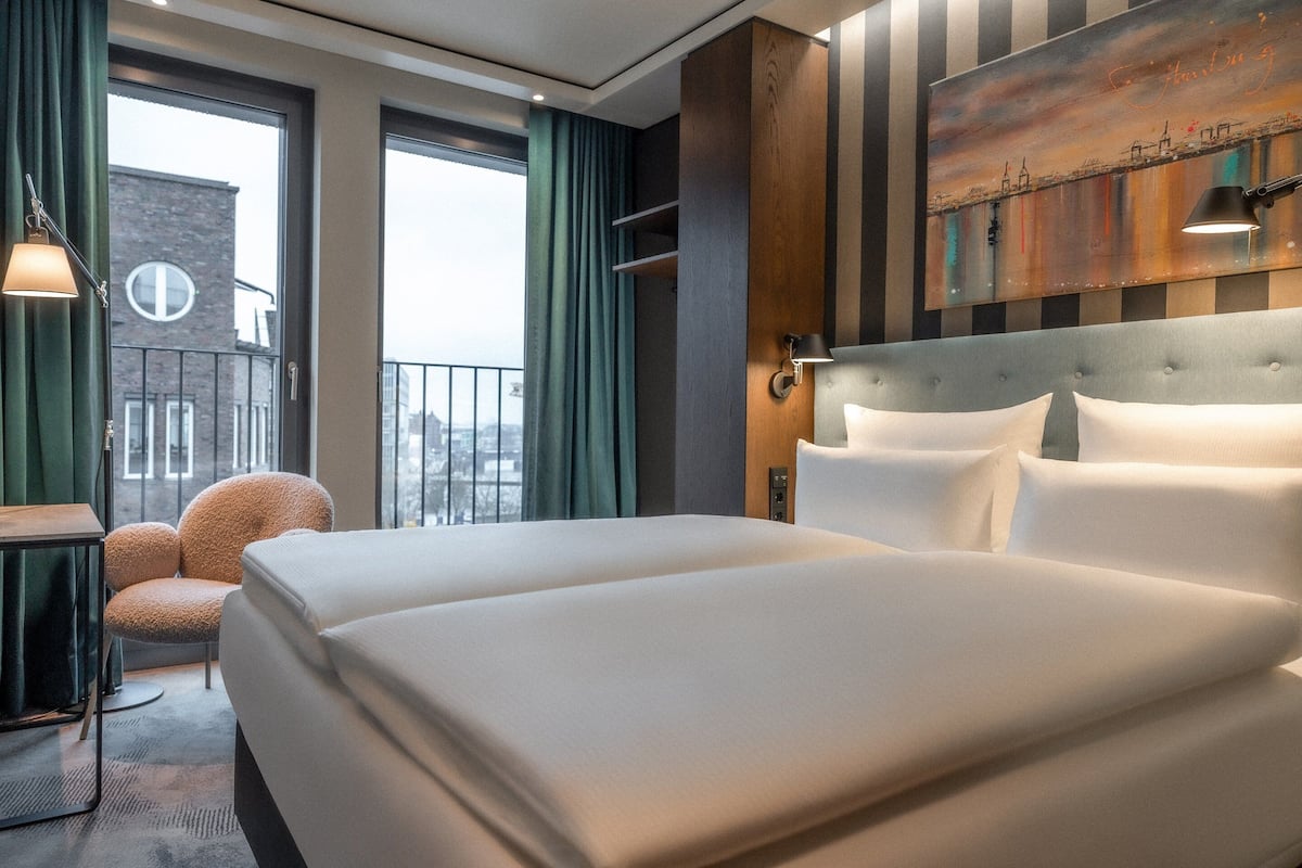 Une chambre d’hôtel avec deux lits, une vue sur la ville, un fauteuil et un tableau au mur.