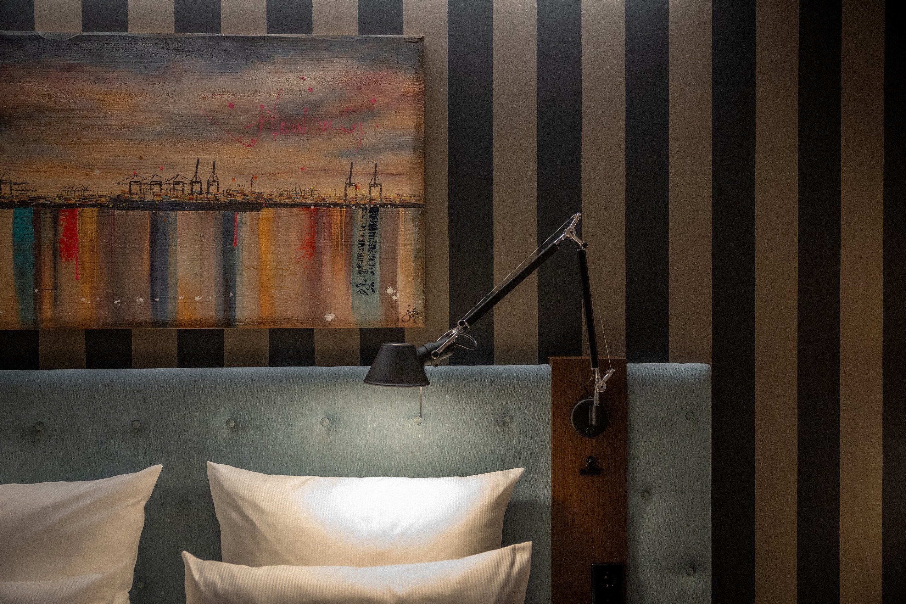 A hotel room with a striped wall, a large painting, a bed with two pillows, and a desk lamp.