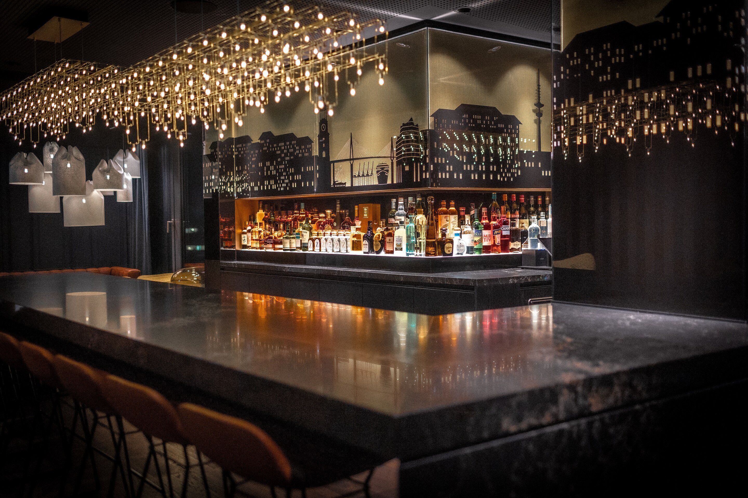 A bar area with a cityscape mural, hanging lights, and a variety of bottles.