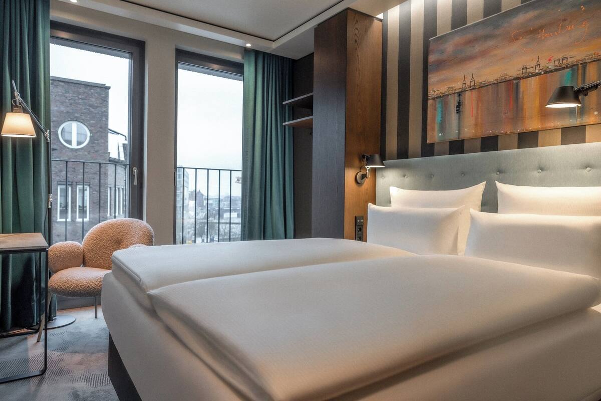 A hotel room with two beds, a view of the city, a chair, and a painting on the wall.