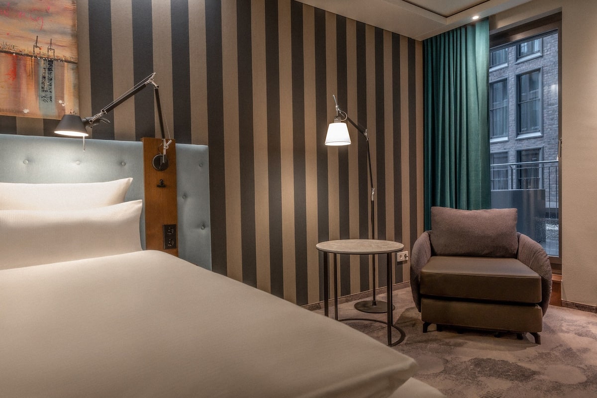 Une chambre d’hôtel avec un mur à rayures, un lit, une chaise, deux lampes et une vue sur un immeuble par la fenêtre.