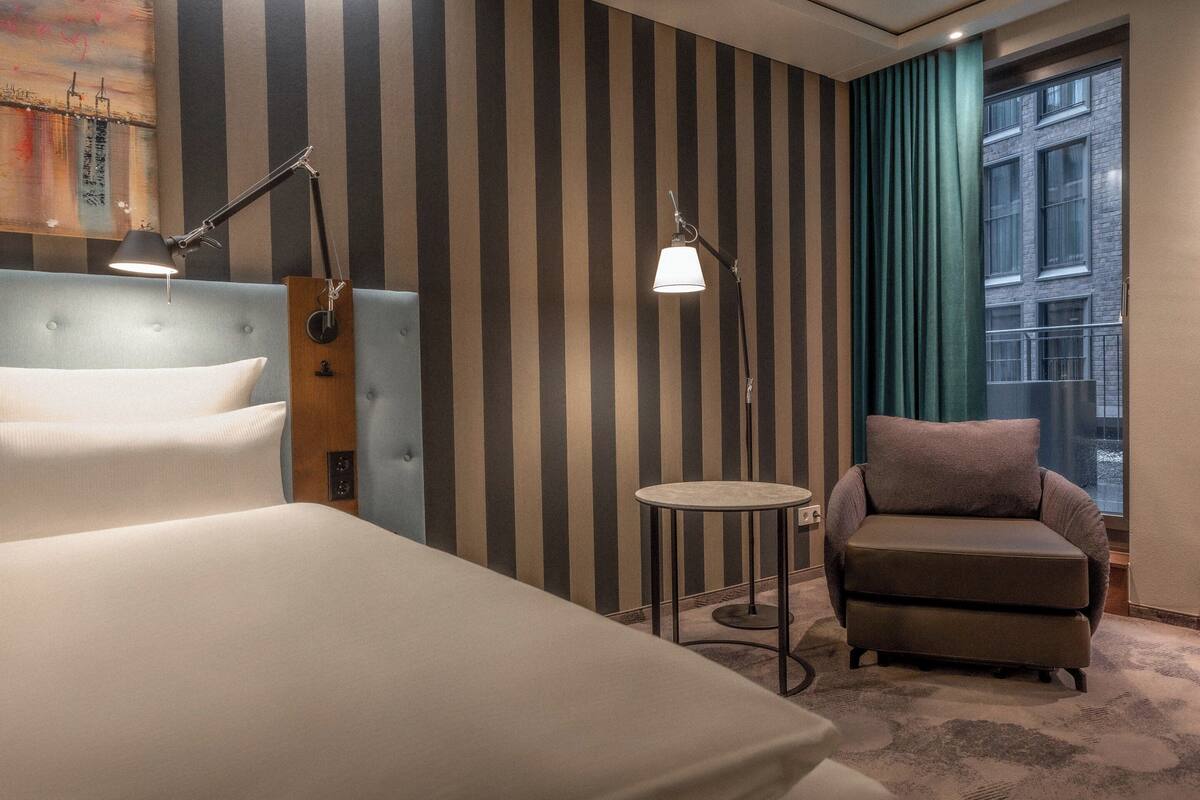 A hotel room with a striped wall, a bed, a chair, two lamps, and a view of a building through a window.