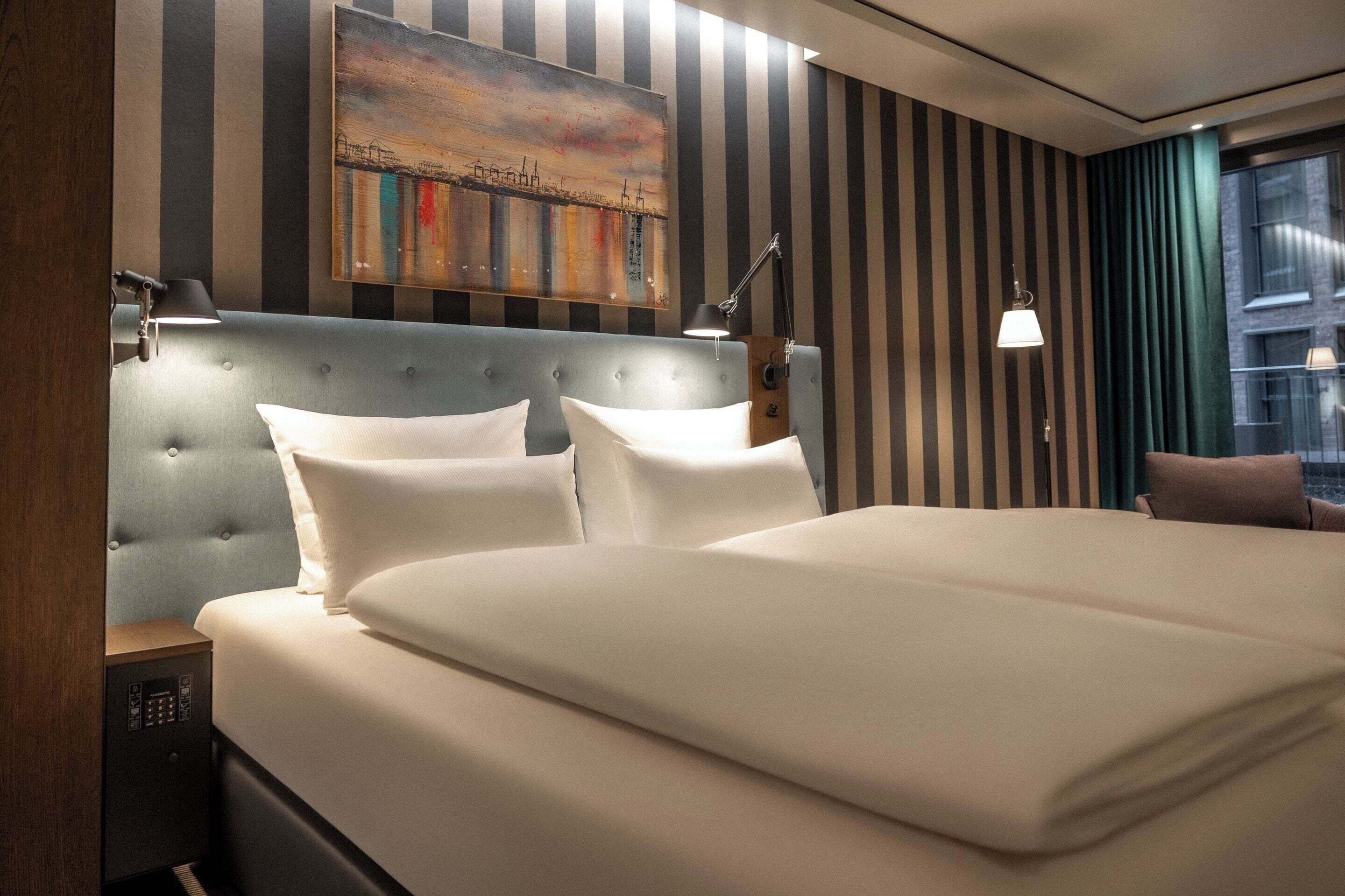 Een hotelkamer met een groot bed, gestreept behang, een schilderij aan de muur en een nachtkastje met een lamp.