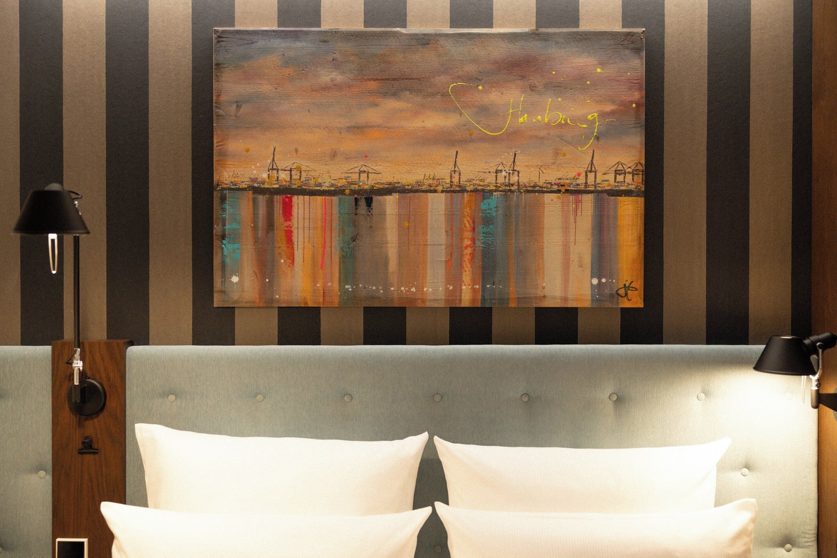 A hotel room with a bed, two bedside lamps, and a cityscape painting on the wall.