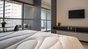Une chambre d’hôtel moderne avec un grand lit, un téléviseur fixé au mur, un balcon offrant une vue sur la ville et une petite table avec des chaises.