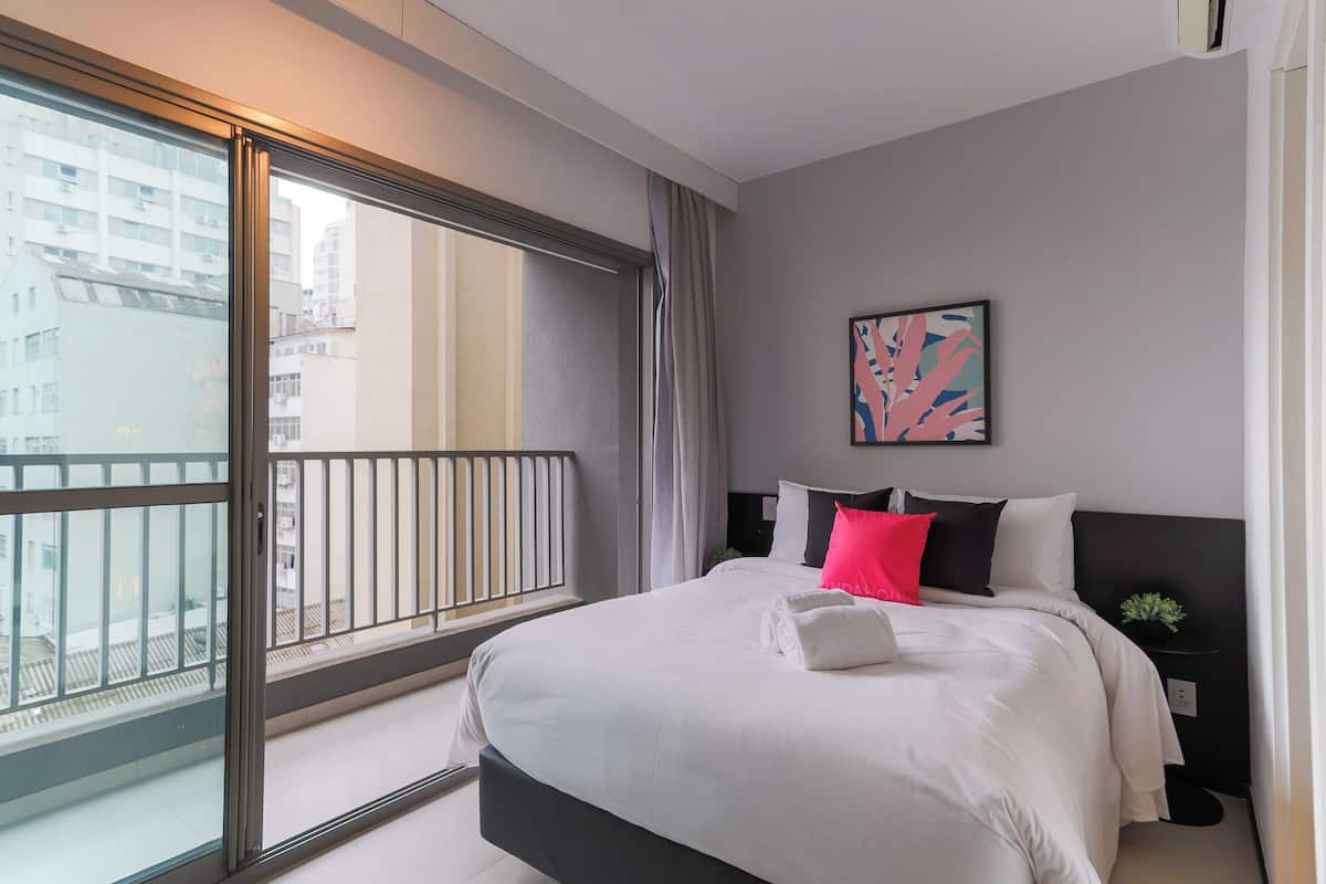 Ein modernes Hotelzimmer mit großem Balkon, einem Bett mit weißer Bettwäsche und einem schwarzen Kopfteil, einem kleinen Nachttisch und einem farbenfrohen abstrakten Gemälde an der Wand.