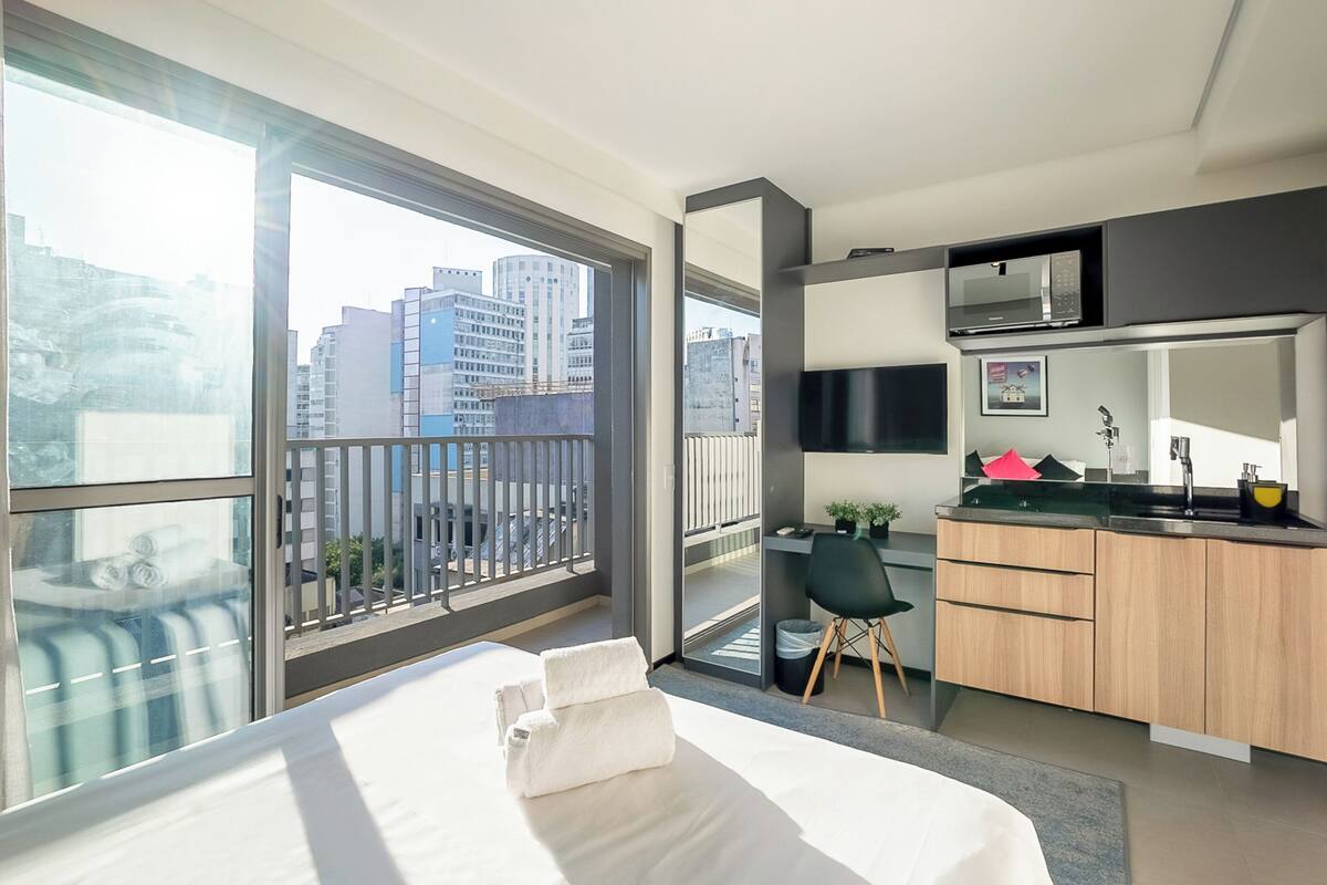 Ein modernes Hotelzimmer mit einem großen Bett, einem kleinen Schreibtisch, einer Küchenzeile und einem Balkon mit Blick auf die Stadt.
