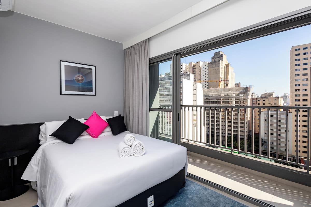 Ein Hotelzimmer mit einem großen Bett, einem gerahmten Bild an der Wand und Blick auf eine Stadtlandschaft vom Balkon aus.