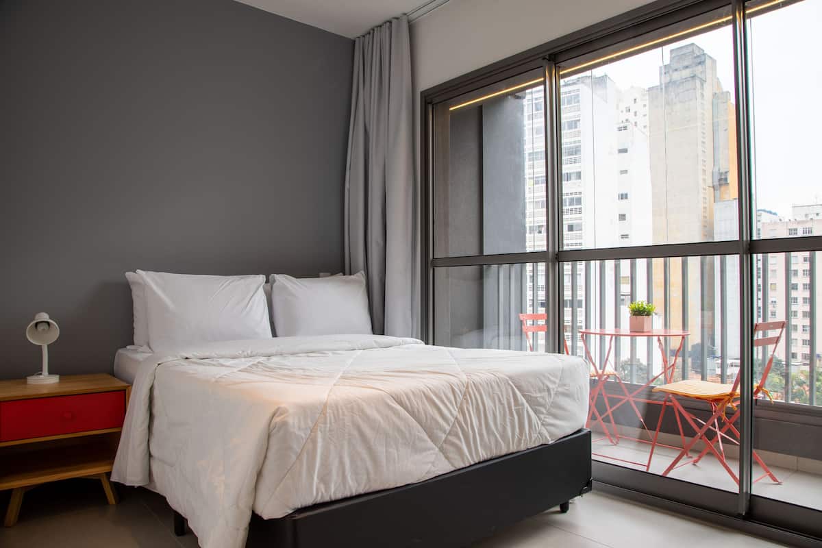 Ein Schlafzimmer mit Bett, Nachttisch, roter Kommode, Fenster mit Blick auf Gebäude und Balkon mit Tisch und Stühlen.