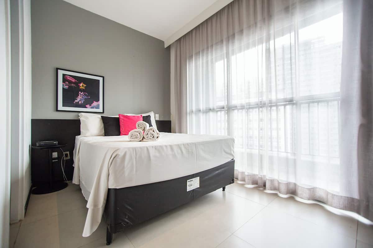 Ein Hotelzimmer mit Bett, Nachttisch, einem gerahmten Bild an der Wand und durchsichtigen Vorhängen am Fenster.
