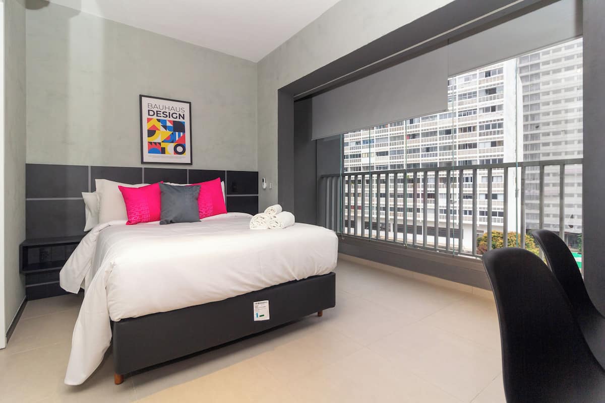 Ein modernes Hotelzimmer mit einem großen Bett, Blick auf die Stadt vom Balkon und einem Bauhaus-Design-Poster an der Wand.