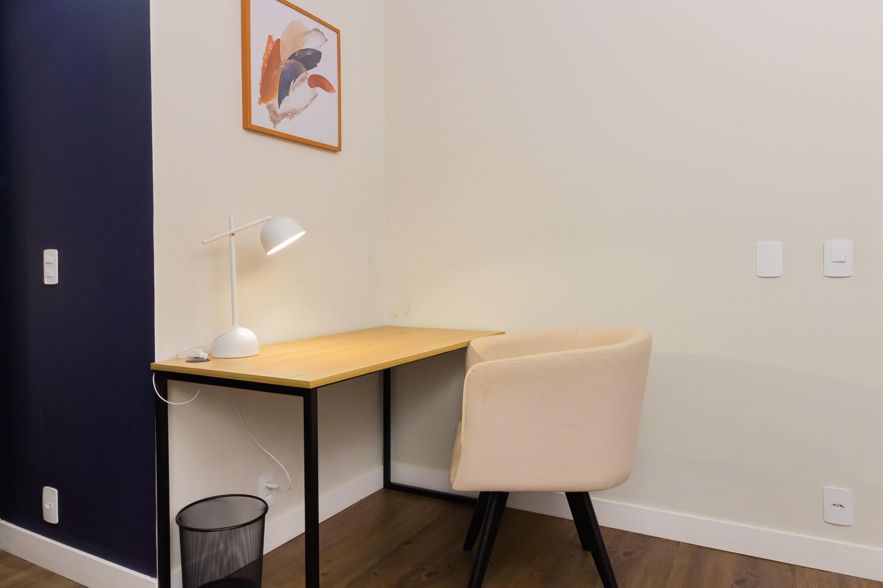 Et hjørneværelse med skrivebord, stol, lampe og et billede på væggen.
