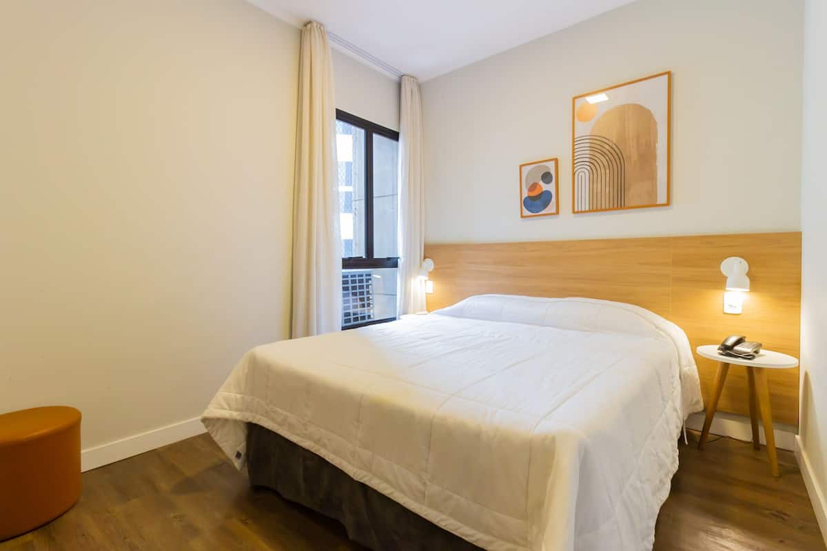 A hotel room with a large bed, a wooden headboard, a small table with a lamp, and two framed artworks on the wall.