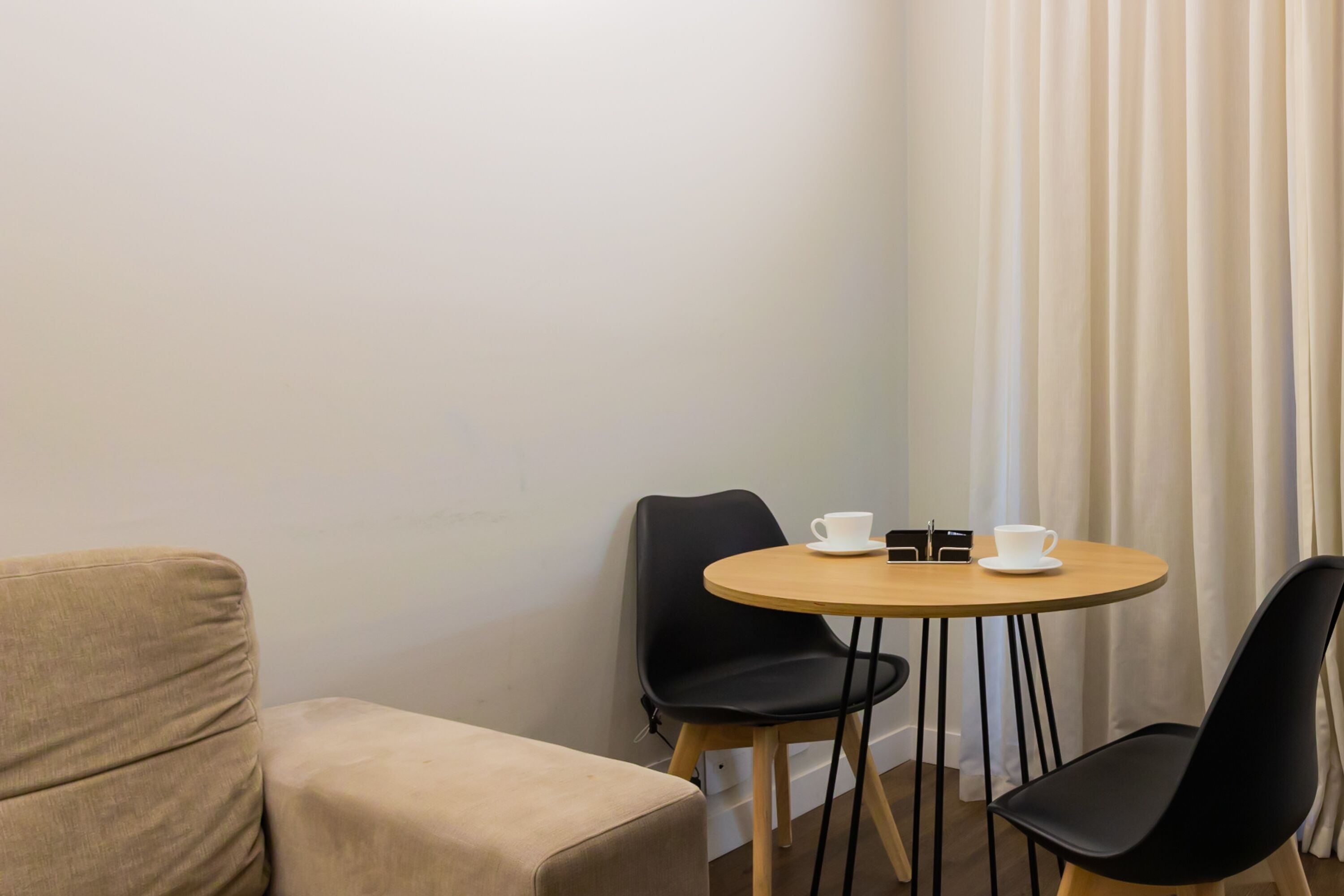 A small, cozy room with a beige sofa, a round wooden table, and two black chairs. There are two white cups on the table.