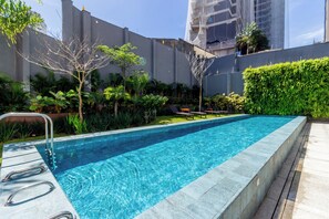 Una piscina rectangular con agua azul cristalina, rodeada por una terraza de madera y un seto bajo.
