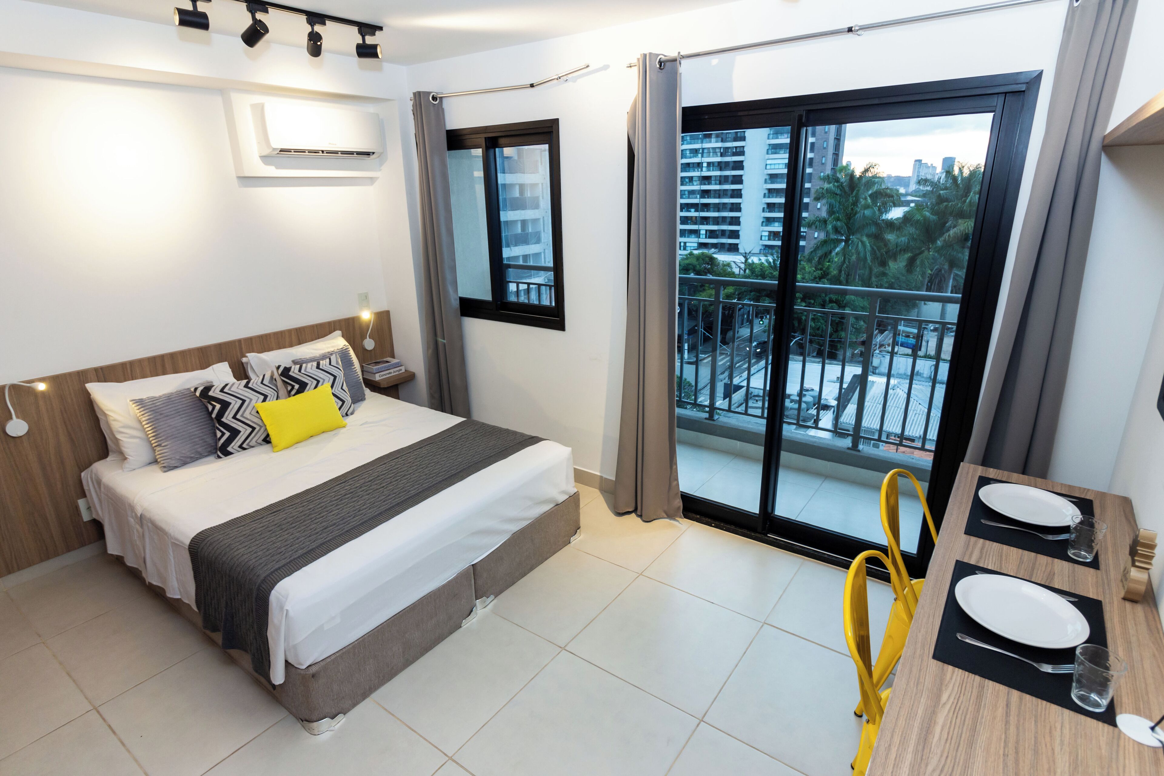 A modern hotel room with a large bed, a balcony with a view of palm trees and buildings, and a dining area with yellow chairs.