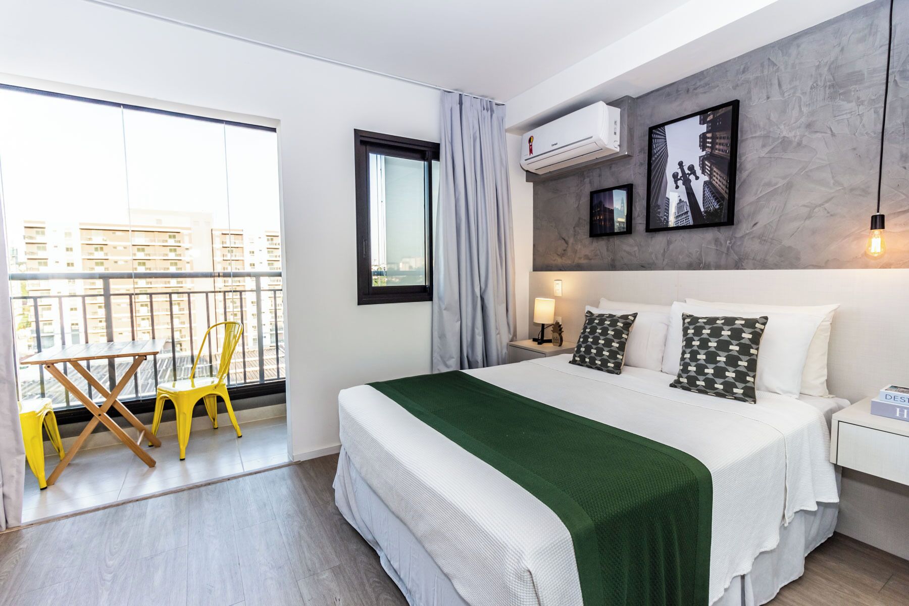 Ein modernes Hotelzimmer mit einem großen Bett, einem Balkon mit gelben Stühlen und Blick auf die Stadt.