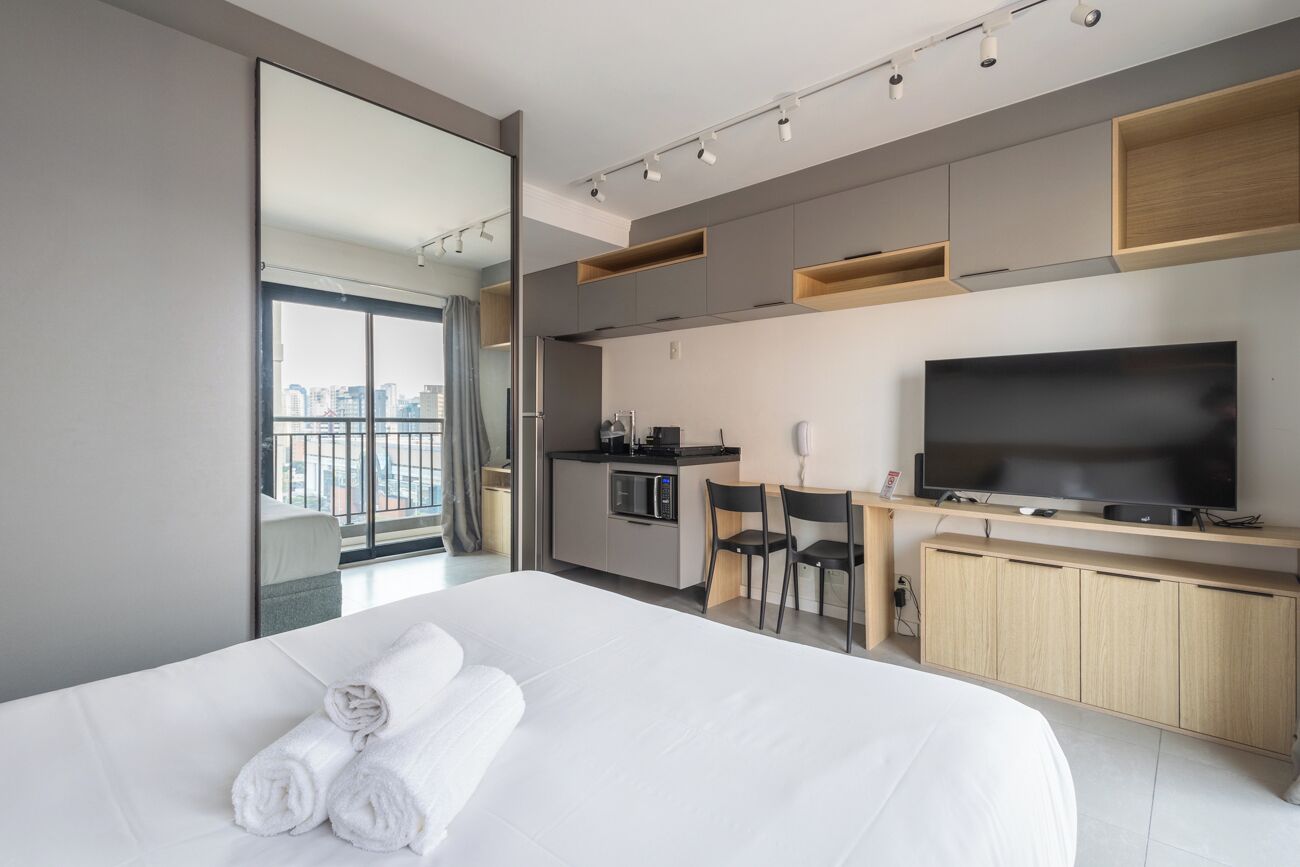 Moderni hotellihuone, jossa on suuri sänky, taulutelevisio, pieni keittiö ja parveke, jolta on näkymä kaupunkiin.