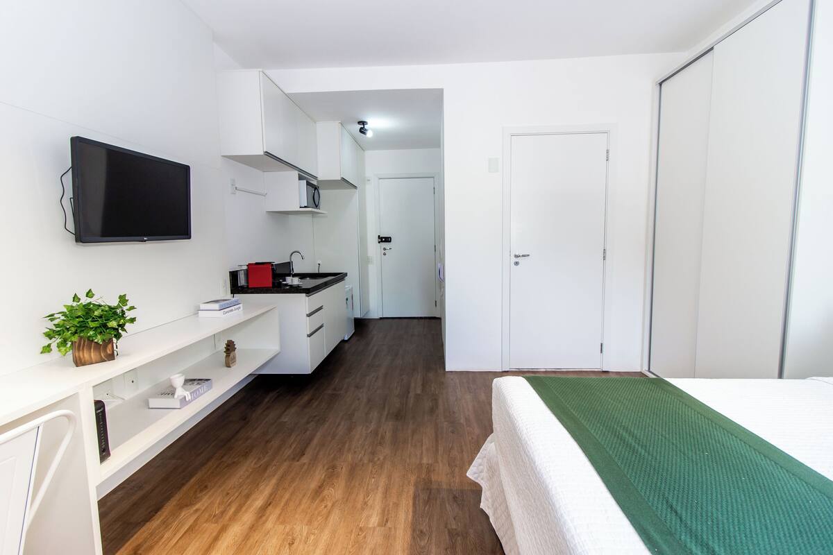 Ein kleines Hotelzimmer mit einem Bett, einem an der Wand befestigten Fernseher, einer Küchenzeile und einer Tür zu einem weiteren Raum.