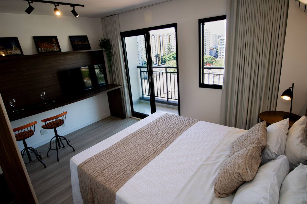 Ein Hotelzimmer mit Bett, Fernseher, einem kleinen Schreibtisch mit zwei Stühlen, einem Balkon mit Blick auf Gebäude und einem fenster mit Vorhängen.