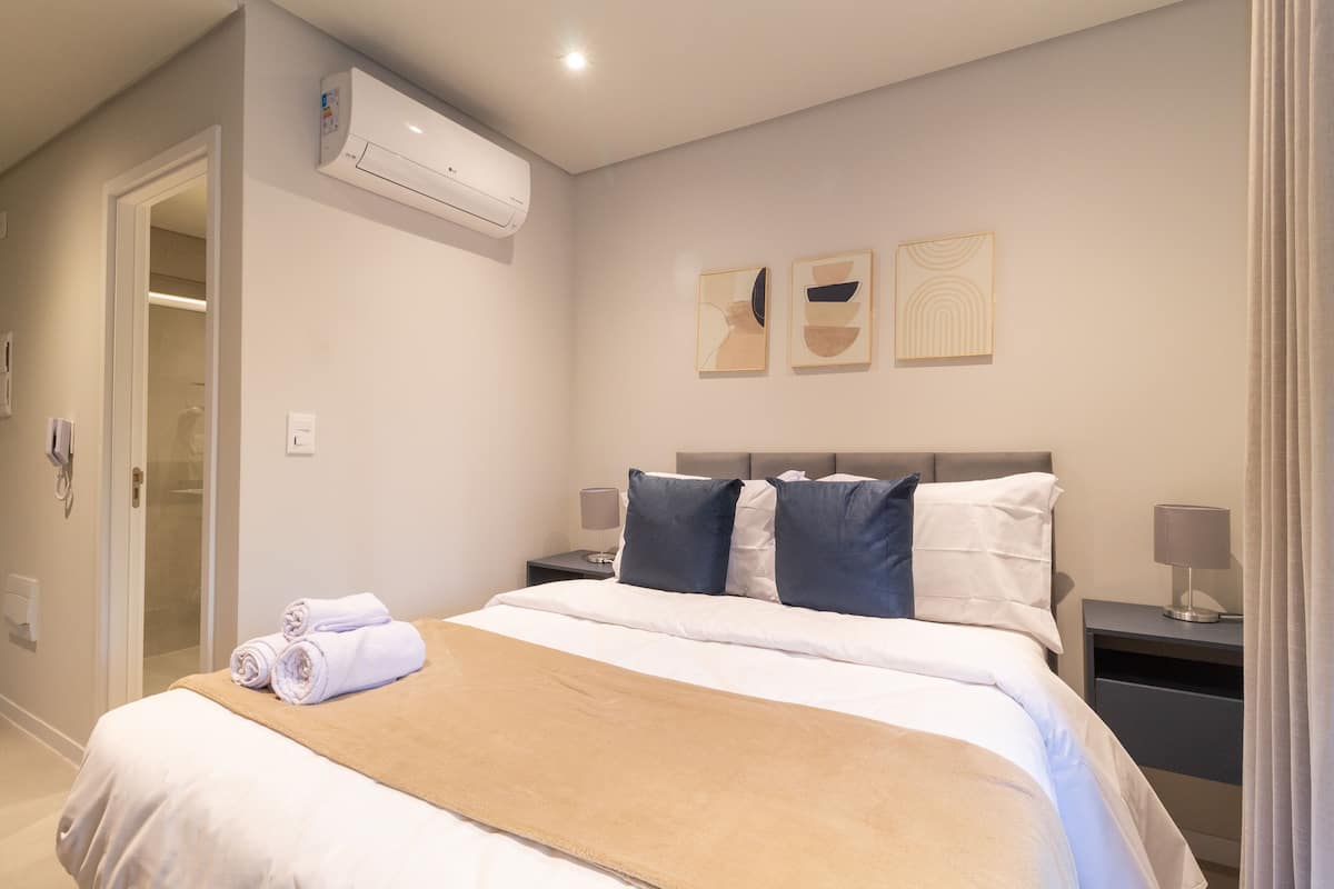 A hotel room with a bed, two bedside tables, an air conditioning unit, and artwork on the wall.