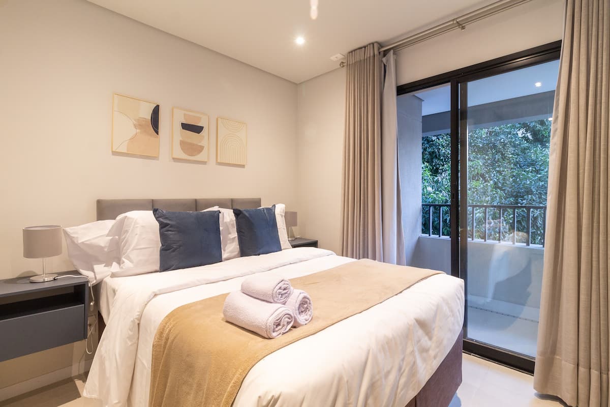 A modern hotel room with a large bed, a nightstand, a lamp, and a view of the outdoors through a sliding glass door.