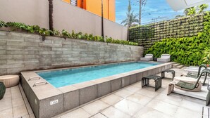 Ein rechteckiger Pool mit Steinbordstein, neben einem Sitzbereich mit Korbnellern und einer Steinmauer mit Grünpflanzen.