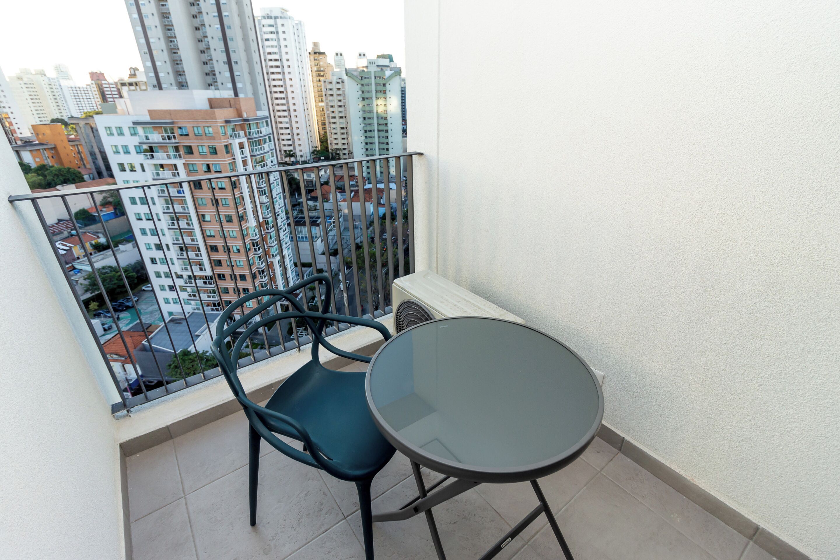 Un balcon avec une table en verre et une chaise, donnant sur un paysage urbain avec de hauts immeubles.