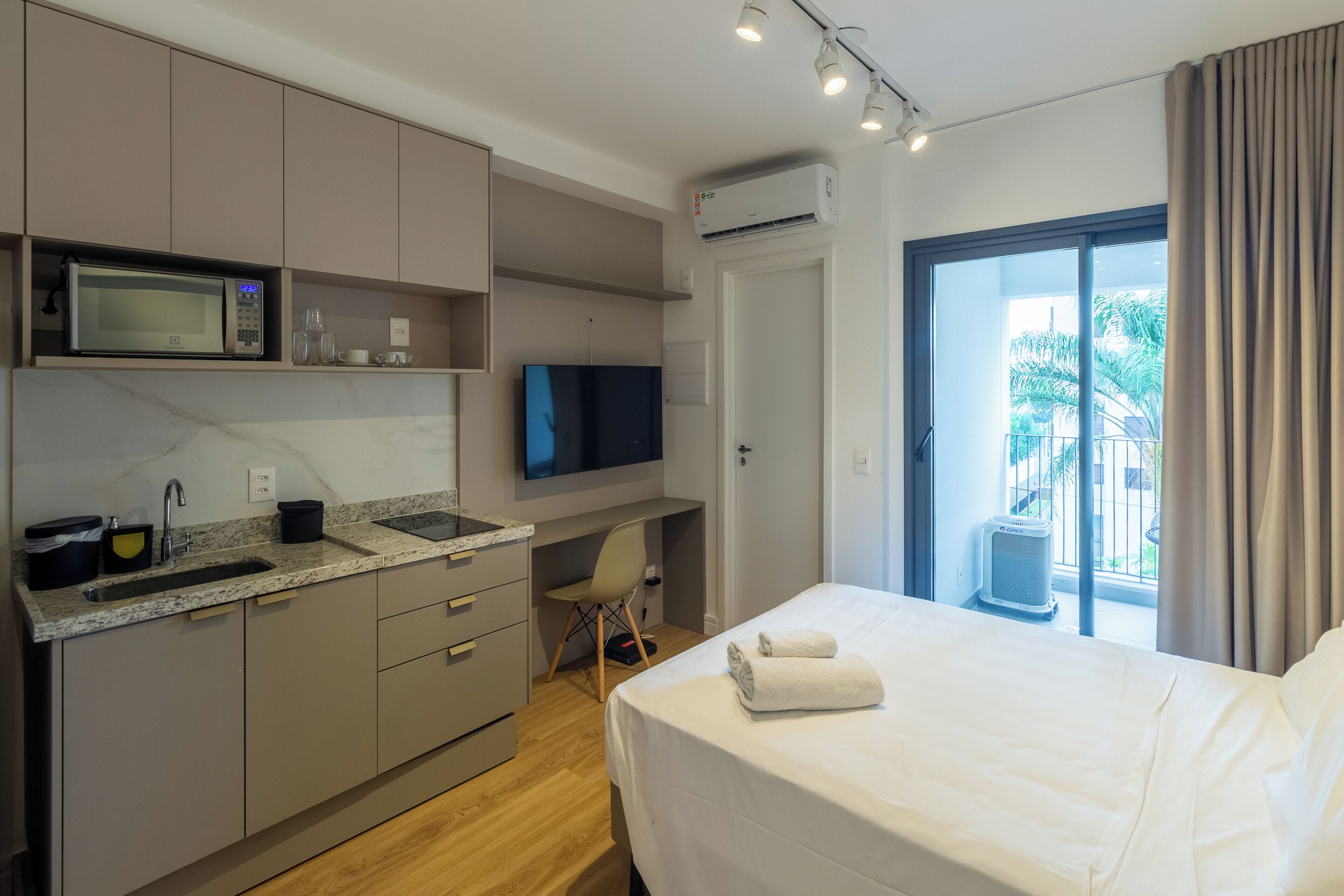 A modern hotel room with a kitchenette, a bed with towels, a TV, and a balcony with curtains.