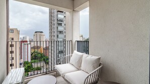 Ein Balkon mit einer weißen, gepolsterten Sitzecke, einem kleinen Tisch und Blick auf hohe Gebäude und Grünflächen.