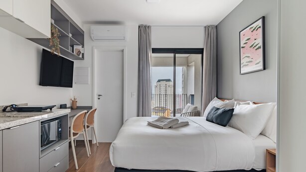 Ein modernes Hotelzimmer mit Bett, einer kleinen Küchenzeile, einem Essbereich und einem Balkon mit Blick auf die Stadt.