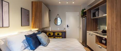 A modern bedroom with a large bed, a small desk, and a built-in kitchen area.