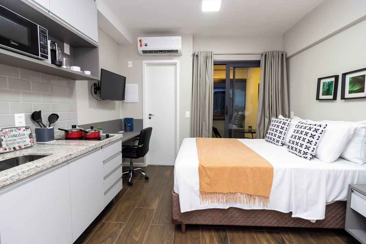 Moderni hotellihuone, jossa on pieni keittiö, valko-oranssilla peitolla varustettu sänky, työpöytä ja televisio.