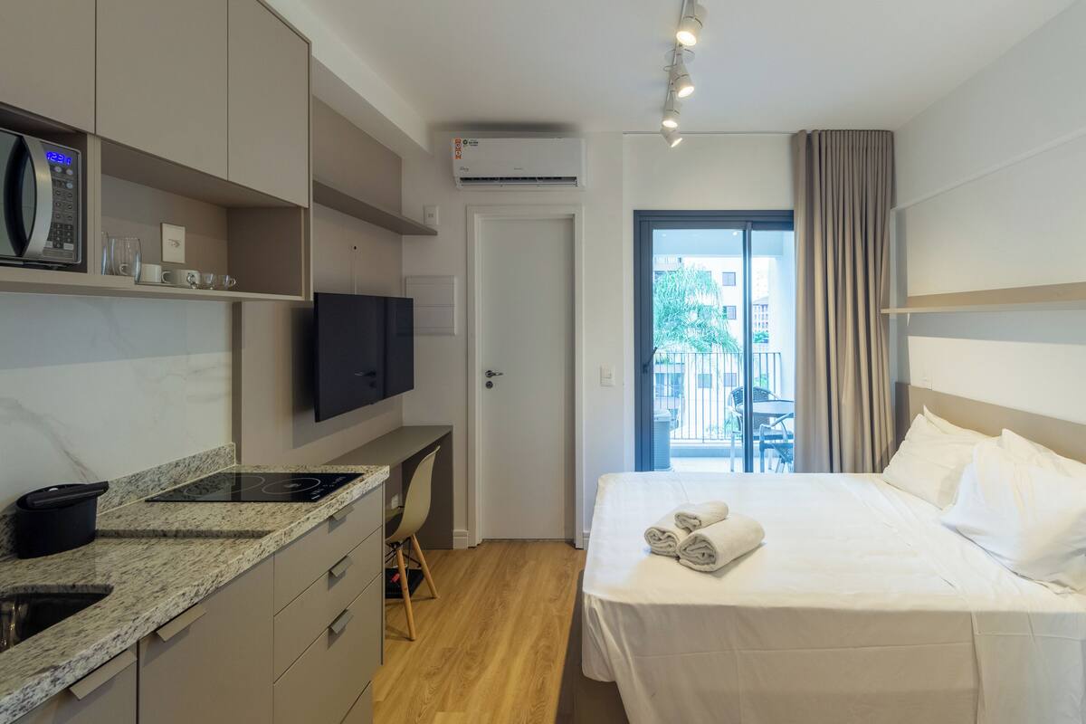 A modern hotel room with a kitchen area, a bed with white linens, and a view of the outdoors.