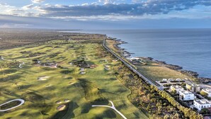 Eine Luftaufnahme eines Küsten-Golfplatzes mit einer langen Straße parallel zum Meer, umgeben von viel Grün und einigen Gebäuden.