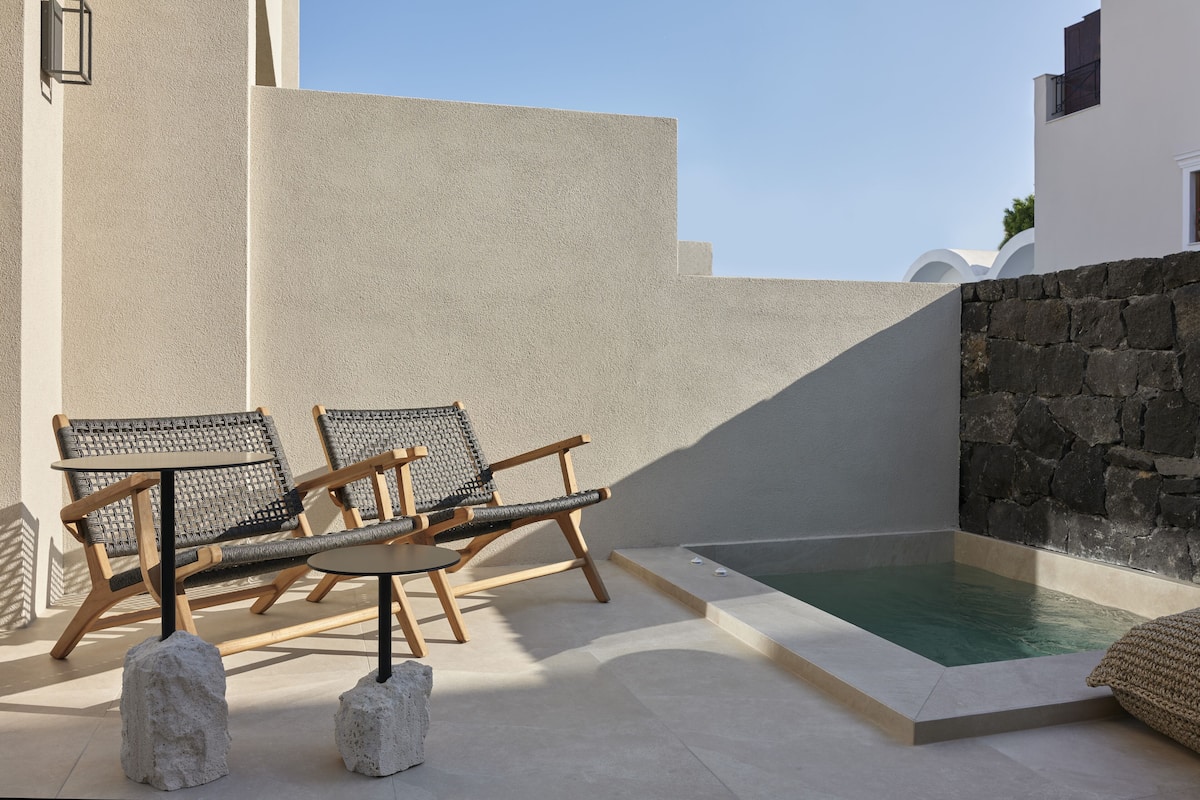 A sunlit outdoor seating area with two wicker chairs and a small round table, a stone wall, and a pool.