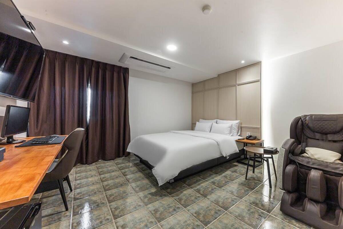 Habitación de hotel con una cama, un escritorio con computadora, una silla de masaje y una mesita con teléfono.