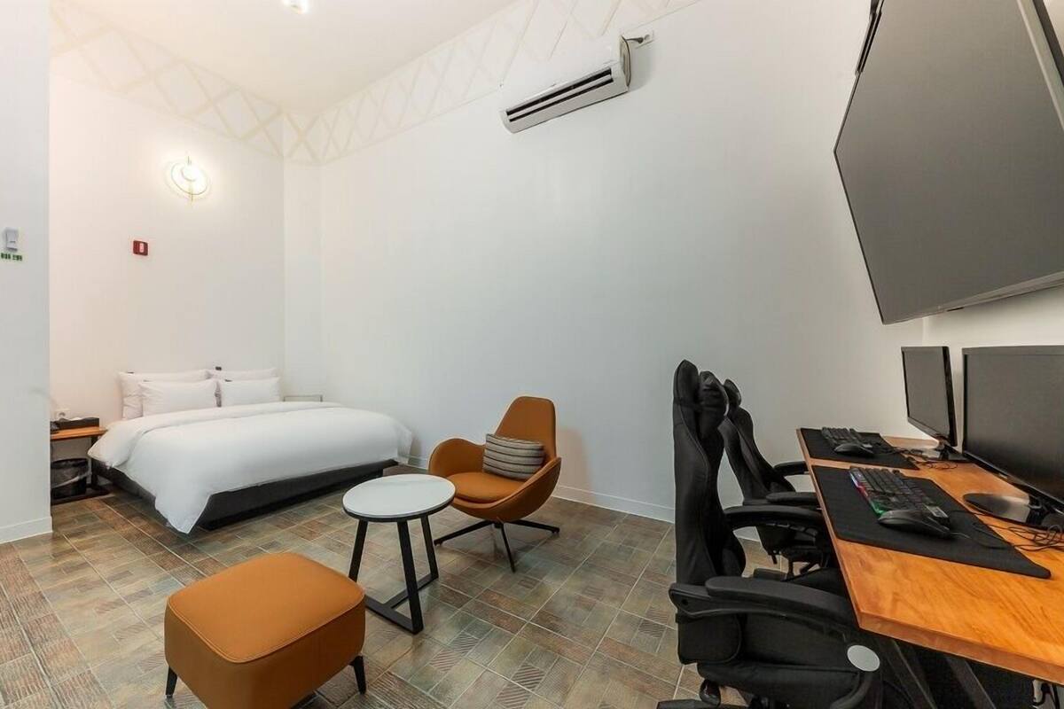 Habitación de hotel con una cama, un escritorio con dos monitores, una silla, una mesita y un equipo de aire acondicionado.