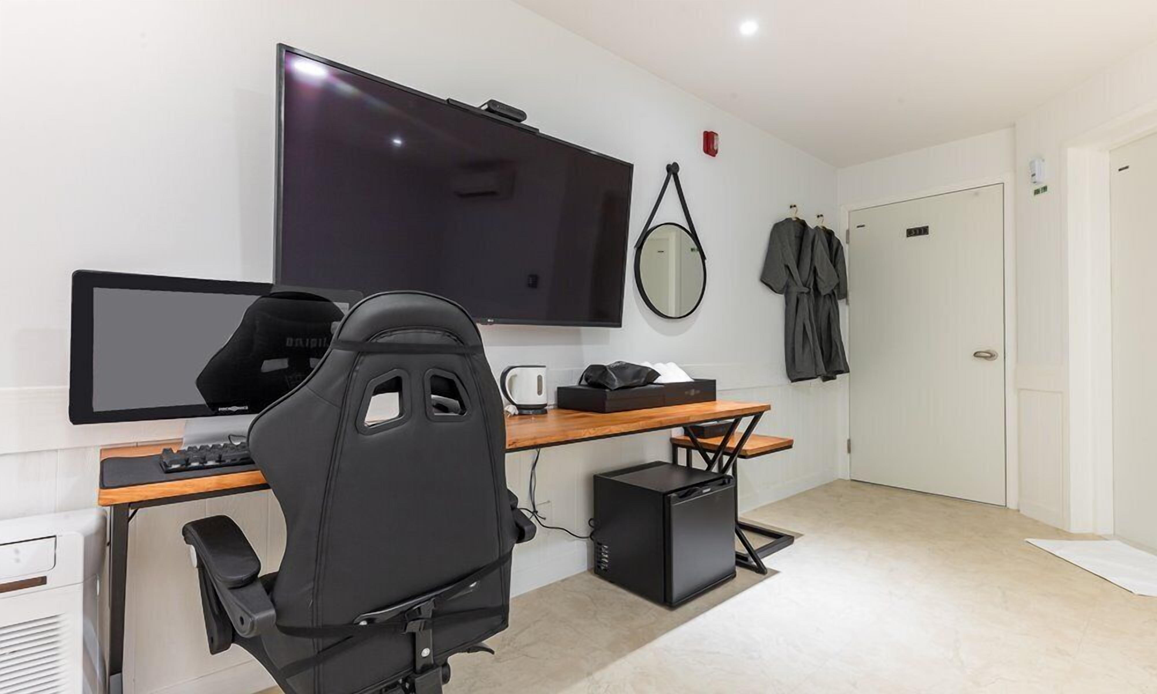 A modern office space with a large flat-screen TV, a gaming chair, a desk with a computer, a coffee maker, and a small black speaker.