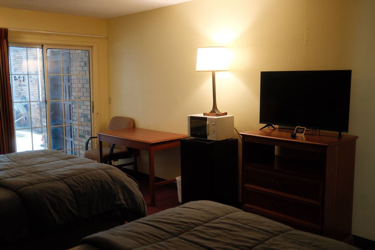 Una habitación de hotel con dos camas, un escritorio con lámpara, televisión y ventana con cortinas.