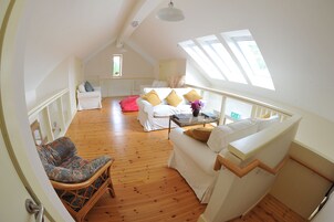 Un espacioso dormitorio tipo loft con techo alto, piso de madera, un sofá blanco, una silla de mimbre y una pequeña mesa con un jarrón de flores.